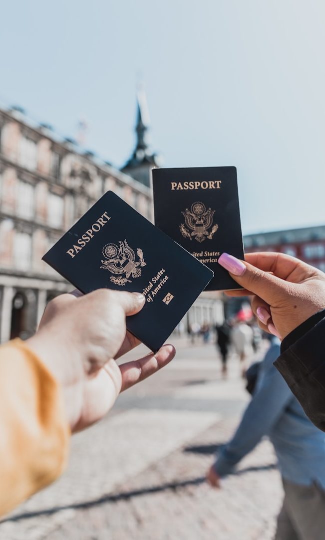 Two hands holding U.S. passports in a plaza with buildings in the background.